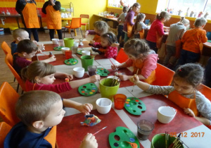 Dzieci siedzą przy stolikach, a niektórzy stoją i ozdabiają swoje figurki ceramiczne.